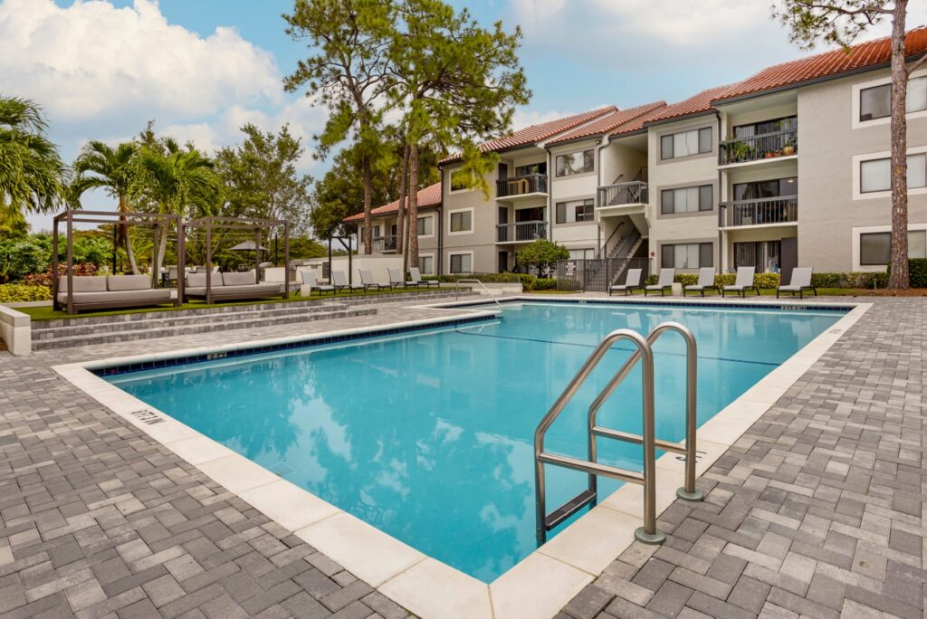 Pool with ladder and steps. Surrounded by lounge chairs and cabanas. Palm trees in the background.