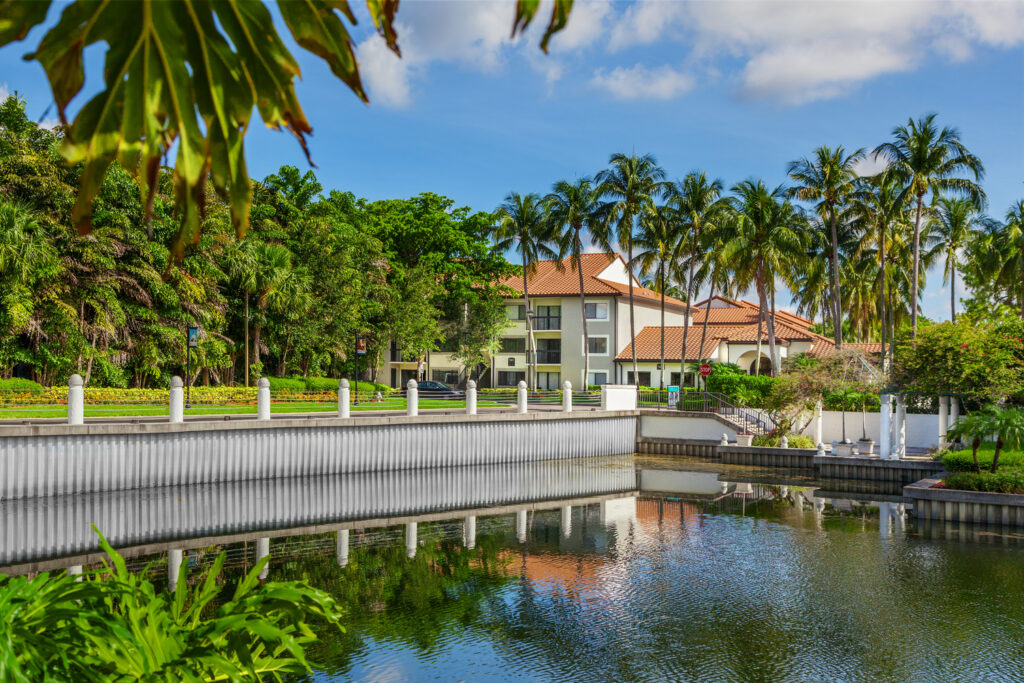 A modern building with a red-tiled roof sits behind a row of palm trees, reflected in a calm pond. A white railing lines the edge of the water, and lush greenery surrounds the scene under a blue sky.