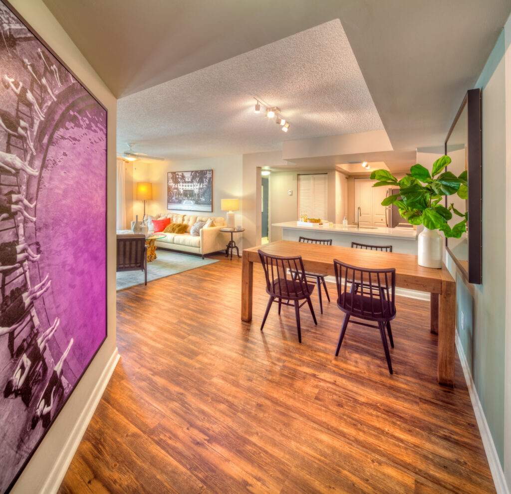 A modern open-concept living and dining area with wood floors, a large wooden dining table with four black chairs, a plant centerpiece, a cozy living room with a sofa and lamps, and large artwork on the left wall.