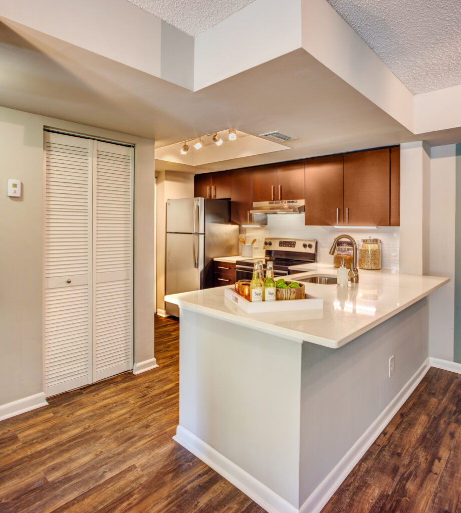 Modern kitchen with brown cabinets, stainless steel appliances, white countertops, and wood flooring. A tray with bottles and glasses sits on the island under track lighting, with a pantry door to the left.