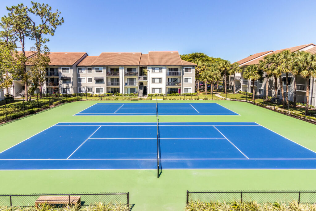 Two blue and green tennis courts outdoors, surrounded by fencing and landscaped greenery, with apartment buildings and palm trees in the background under a clear blue sky.